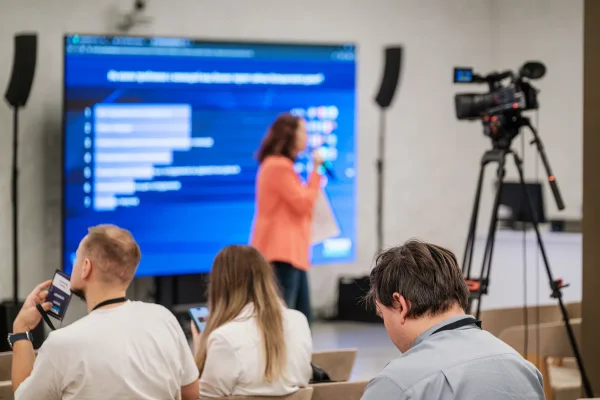 Spreker op locatie voor een groot scherm met live resultaten, vastgelegd door een camera op statief voor de online kijkers.