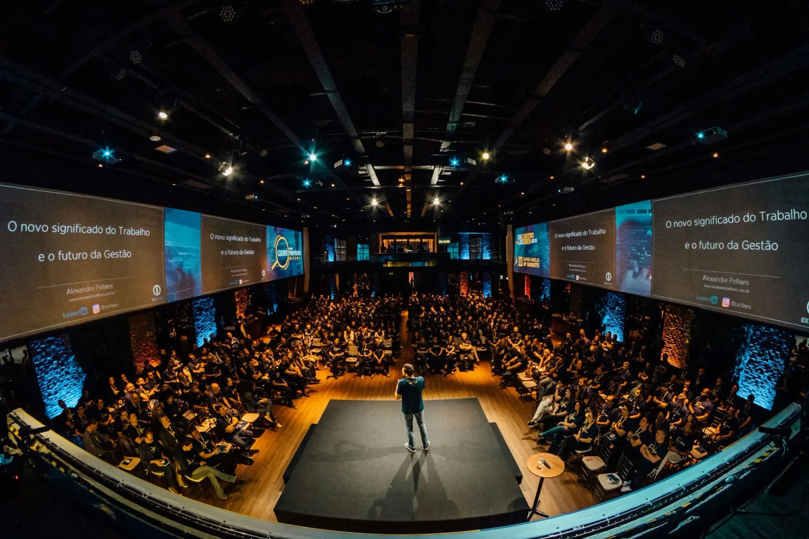 Volle zaal met een spreker op een rond podium en panoramische schermen, tijdens een hybride event.