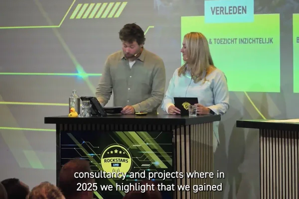 Presentatieduo achter een desk in een studiodecor, met ondertiteling in beeld voor het internationale publiek.