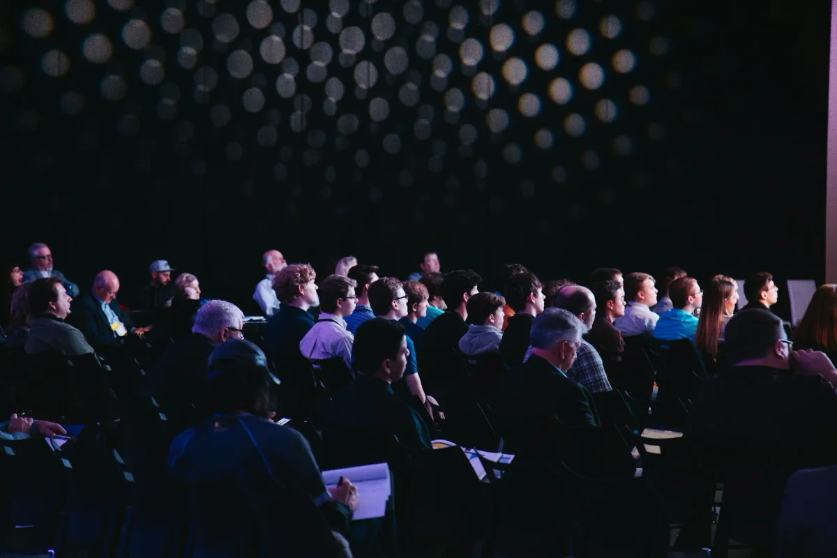 Publiek in een verduisterde zaal kijkt naar het podium tijdens een live event.
