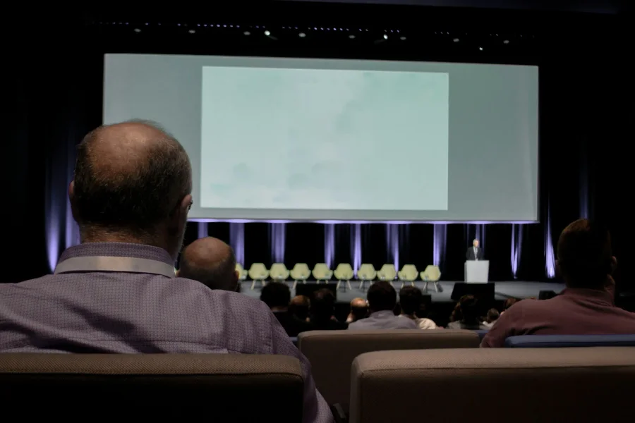 Publiek in de zaal kijkt naar het podium met sprekers en een groot scherm, terwijl de sessie ook online wordt uitgezonden.