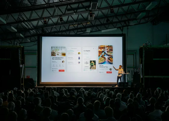 Spreker op een podium voor een groot scherm in een volle zaal tijdens de introductie van een nieuw product.